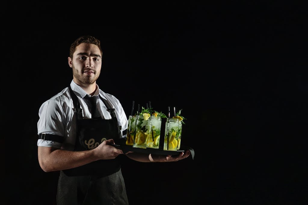 A smiling waiter in uniform holds a tray with refreshing lemon drinks against a black background.